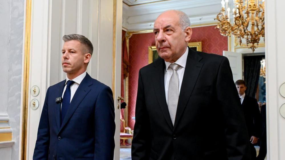 Peter Magyar and Tamas Sulyok walking through a door. A chandelier hags in the room behind them, which also has a room with red-coloured wallpaper.