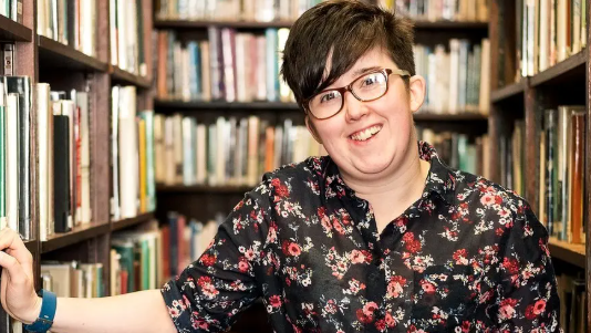 Lyra McKee is wearing a black and red floral shirt. She is standing in front of bookcases in a library. Her hand is rested on a shelf and she is smiling.