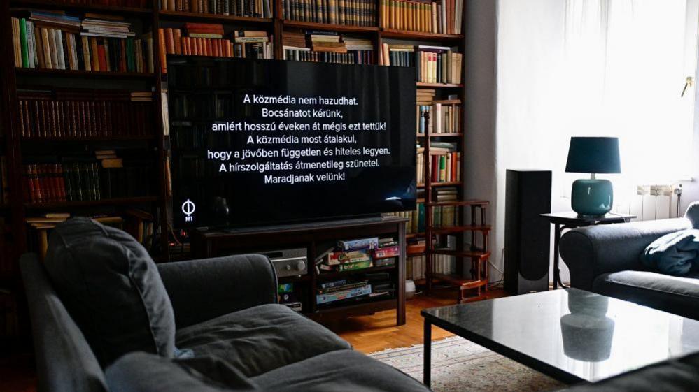 A TV screen is seen inside a living room in front of a large bookcase and behind a coffee table and two sofas. The screen is black with white writing that reads "State media cannot lie. We apologise for having done so for years".
