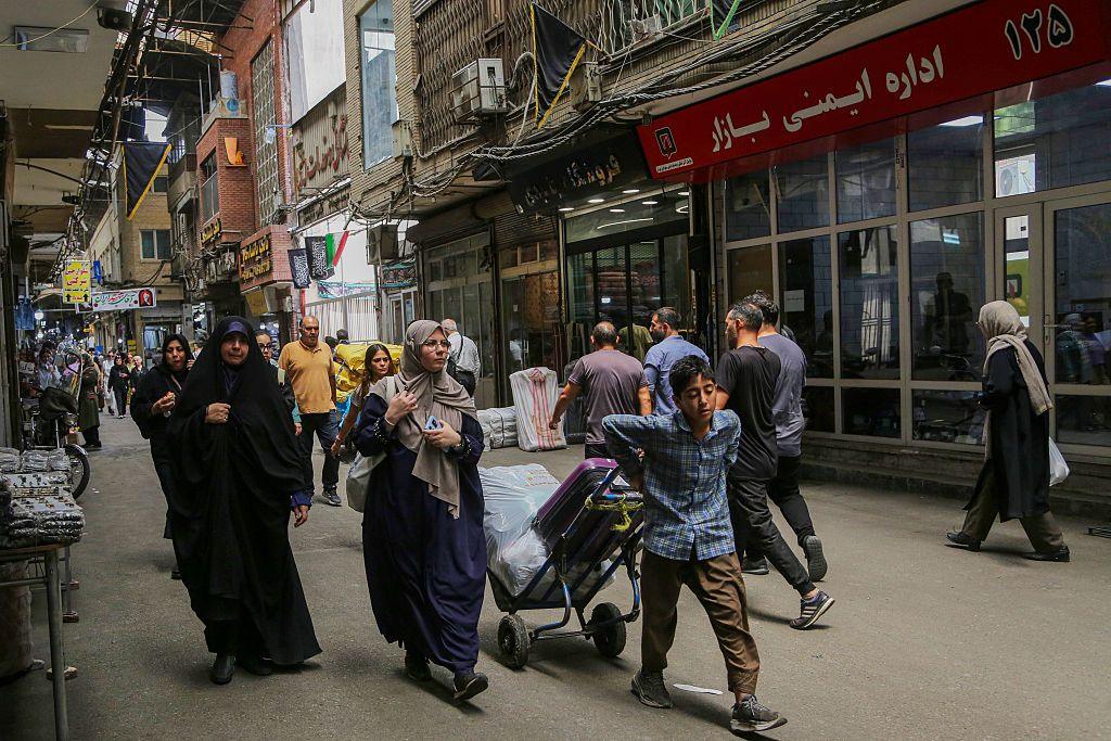 Wide shot showing a busy street with shops in Tehran. There are several men and women walking through the street At the front of the image is a young boy wheeling a trolley behind him.