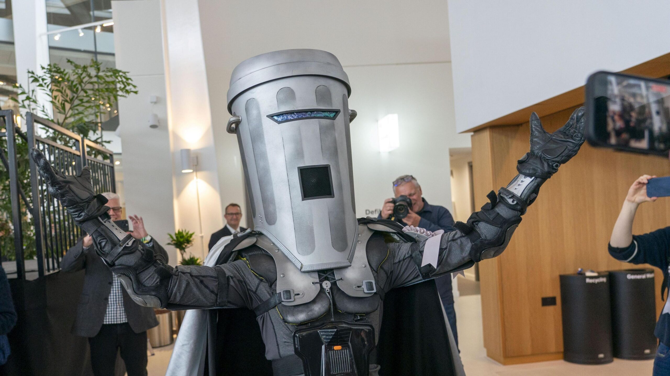 Count Binface - a man dressed in a grey space costume with a cape on the back and a dustbin for the head. He has his arms outstretched in the air while inside a venue, with a number of photographers taking pictures of him.
