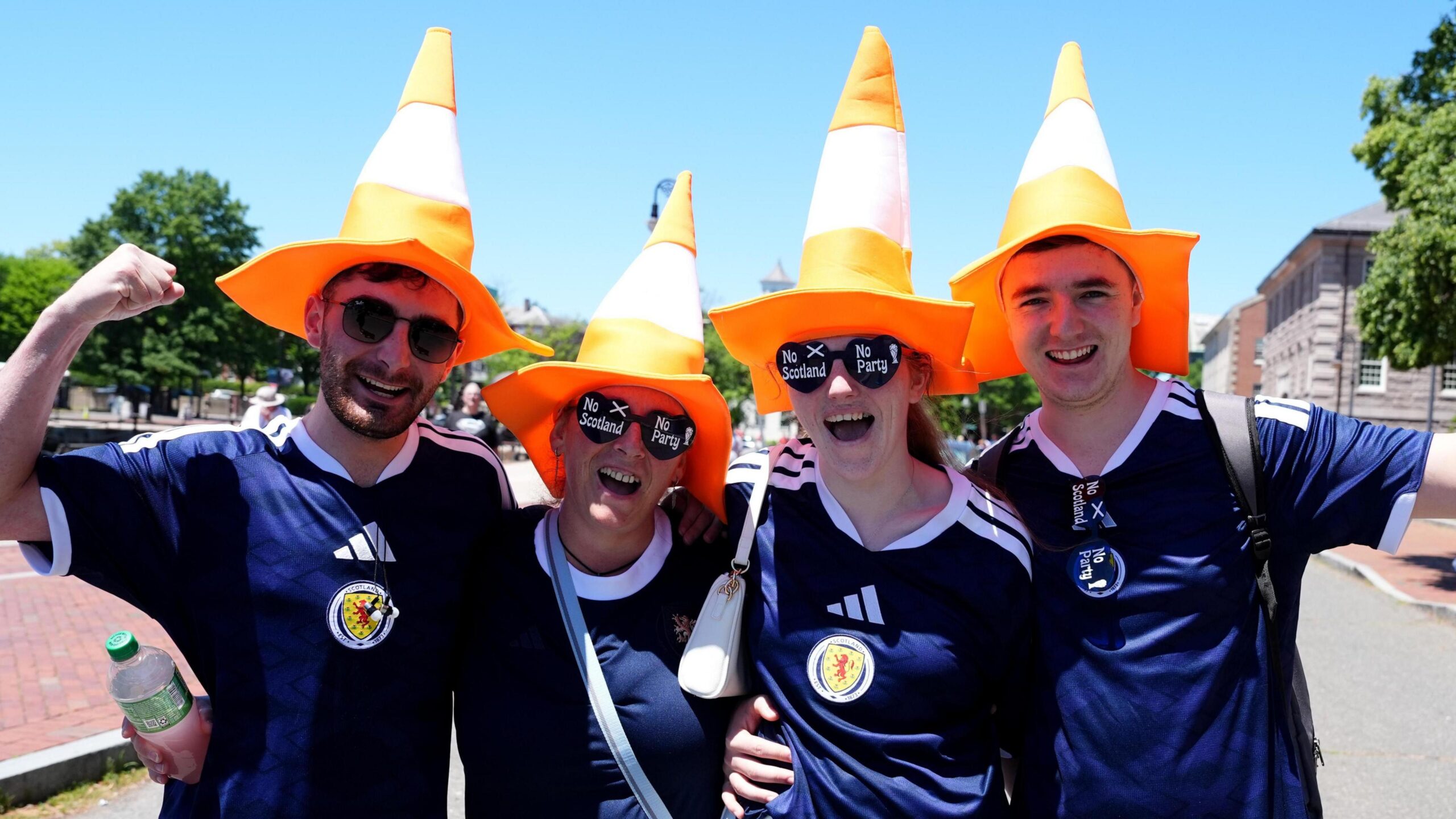 Four Scotland football fans wearing blue Scotland football shirts and orange traffic cone hats. They are cheering towards the camera.