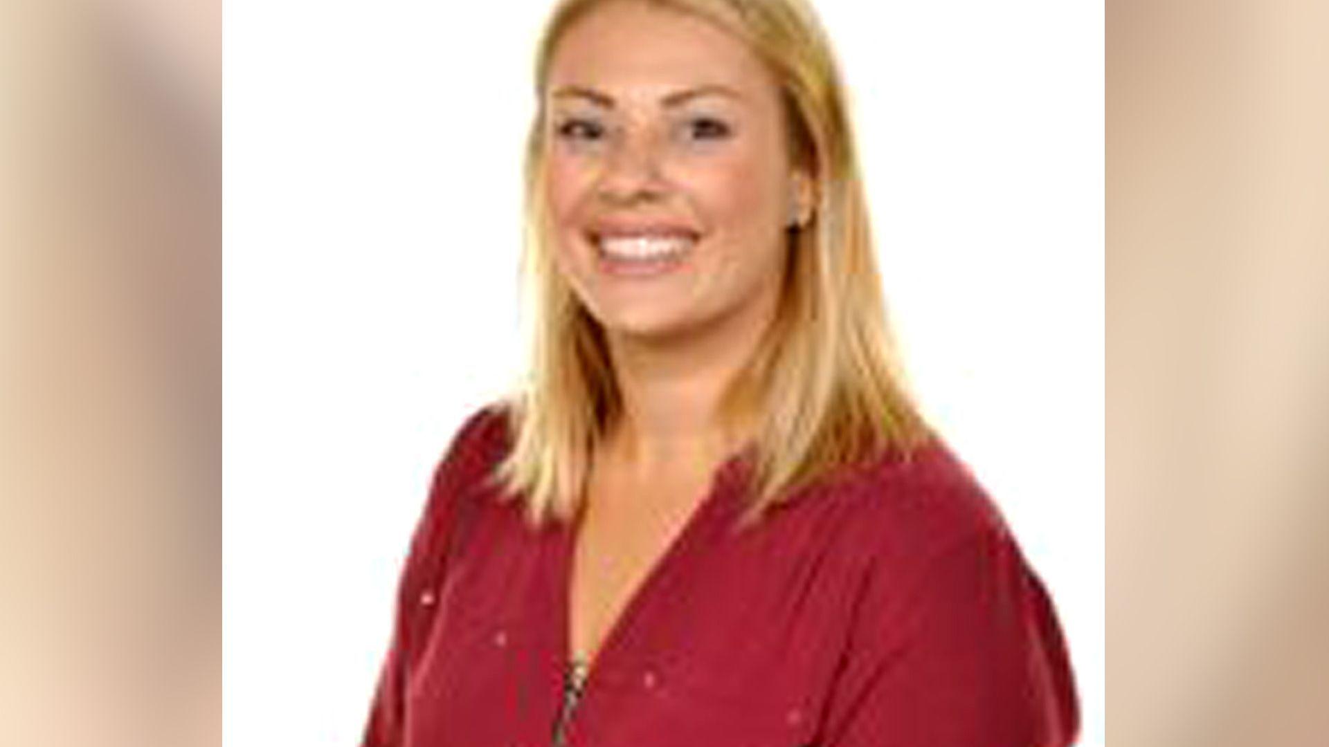 A woman smiling at the camera. It is a head and shoulders shot. She has blonde hair and a red top on.