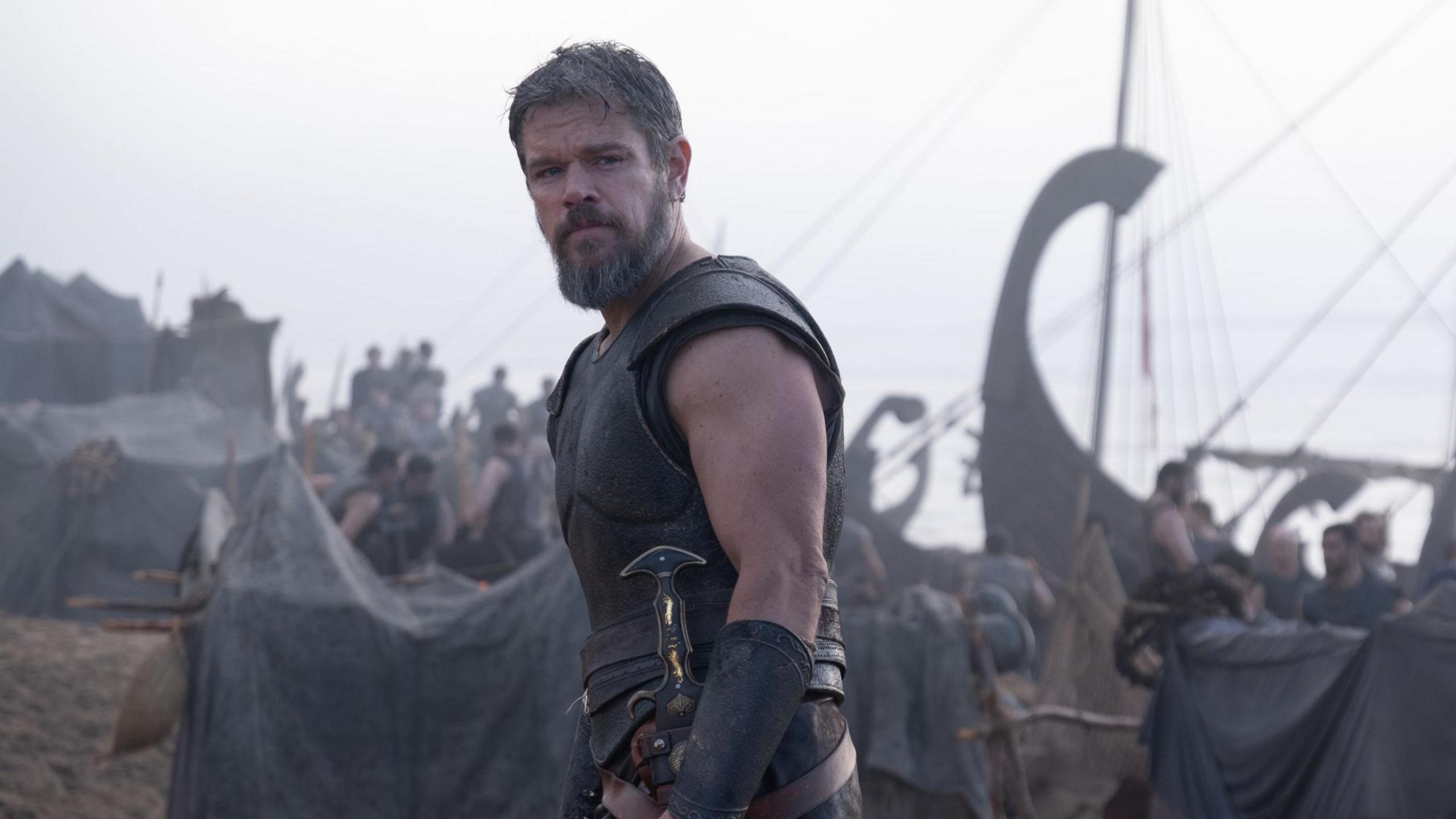 Matt Damon looking to camera in armour, ships and warriors in background