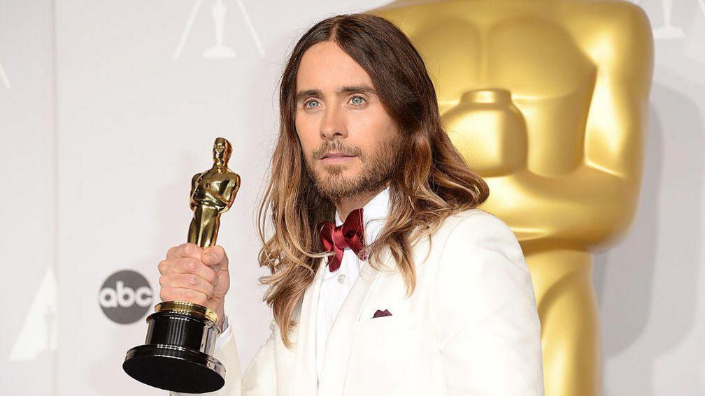 Jared Leto in 2014, wearing a white suit and satin red bow tie, poses for cameras holding up his Oscar award with one hand. He is against a white background and there is a large gold replica of the award behind him.