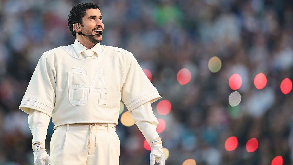 A man dressed all in white - trousers, shirt and tie, gloves, and with a cropped American Football shirt on top. He has a microphone attached to his face and there is an out-of-focus crowd in the background