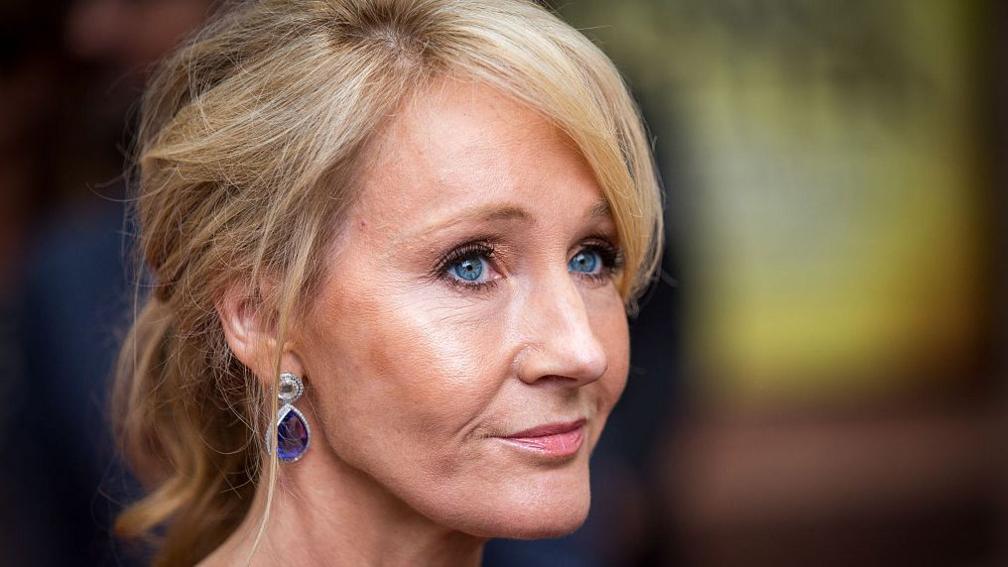 A woman - JK Rowling- with blonde hair, wearing tear drop shaped earrins, viewed side on