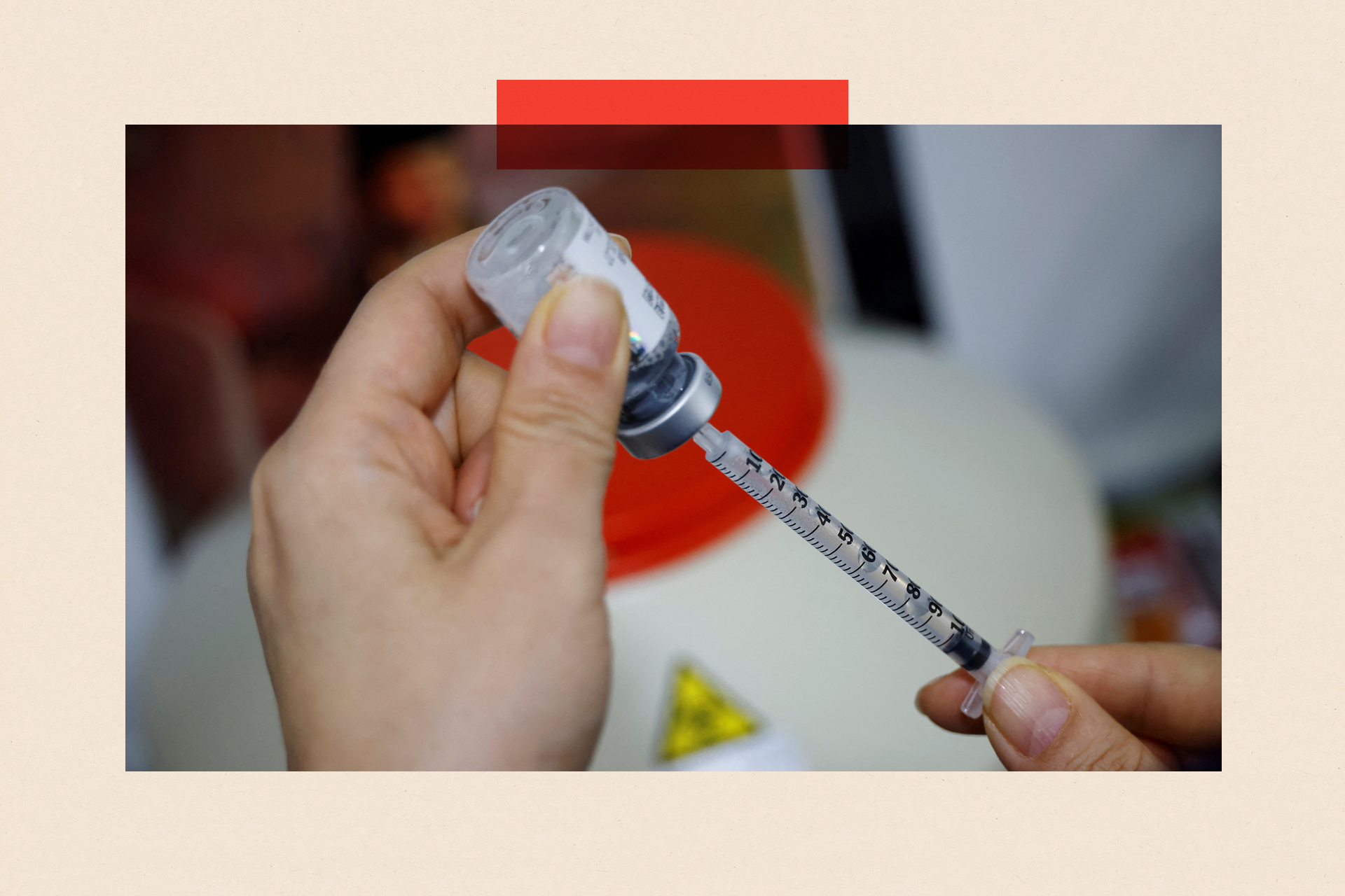 The hands of a nurse prepare a Botox injection by drawing liquid from a container using a syringe. In the background is a much larger container and a triangular yellow and black toxic sign appears out of focus.