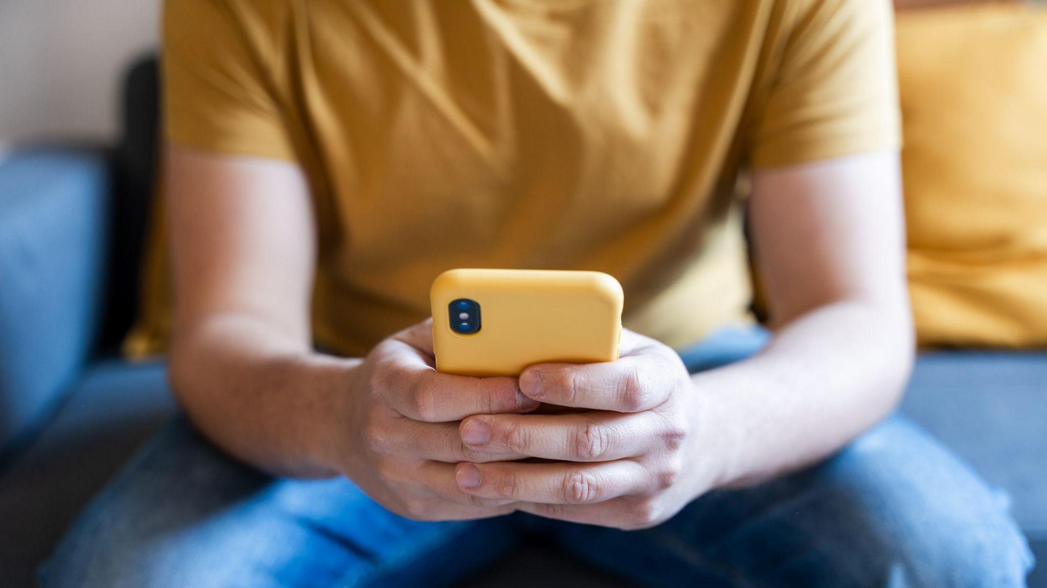 A close up of a person holding a yellow phone, sitting on a blue sofa, wearing jeans and a yellow t-shirt.