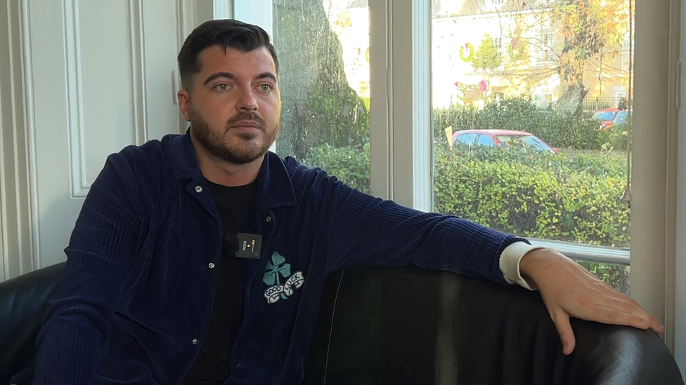 Luke Worley sat on a black leather sofa while listening to a reporter, who is off-camera to the right. He has short black hair and a beard, and is wearing a navy blue jacket over a black T-shirt. Behind him is a window, and there is a hedge and the top of a red car outside.