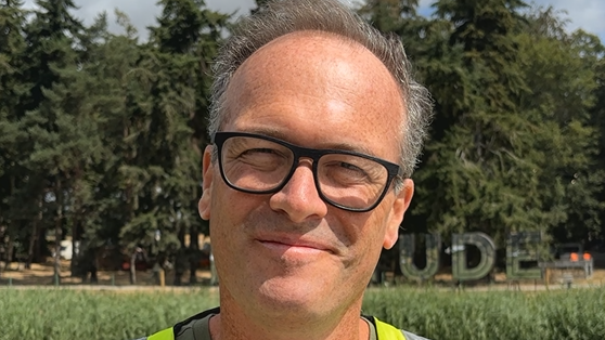 A head shot of a man wearing glasses. He is standing in a field with a mirrored Latitude sign behind him in the distance.