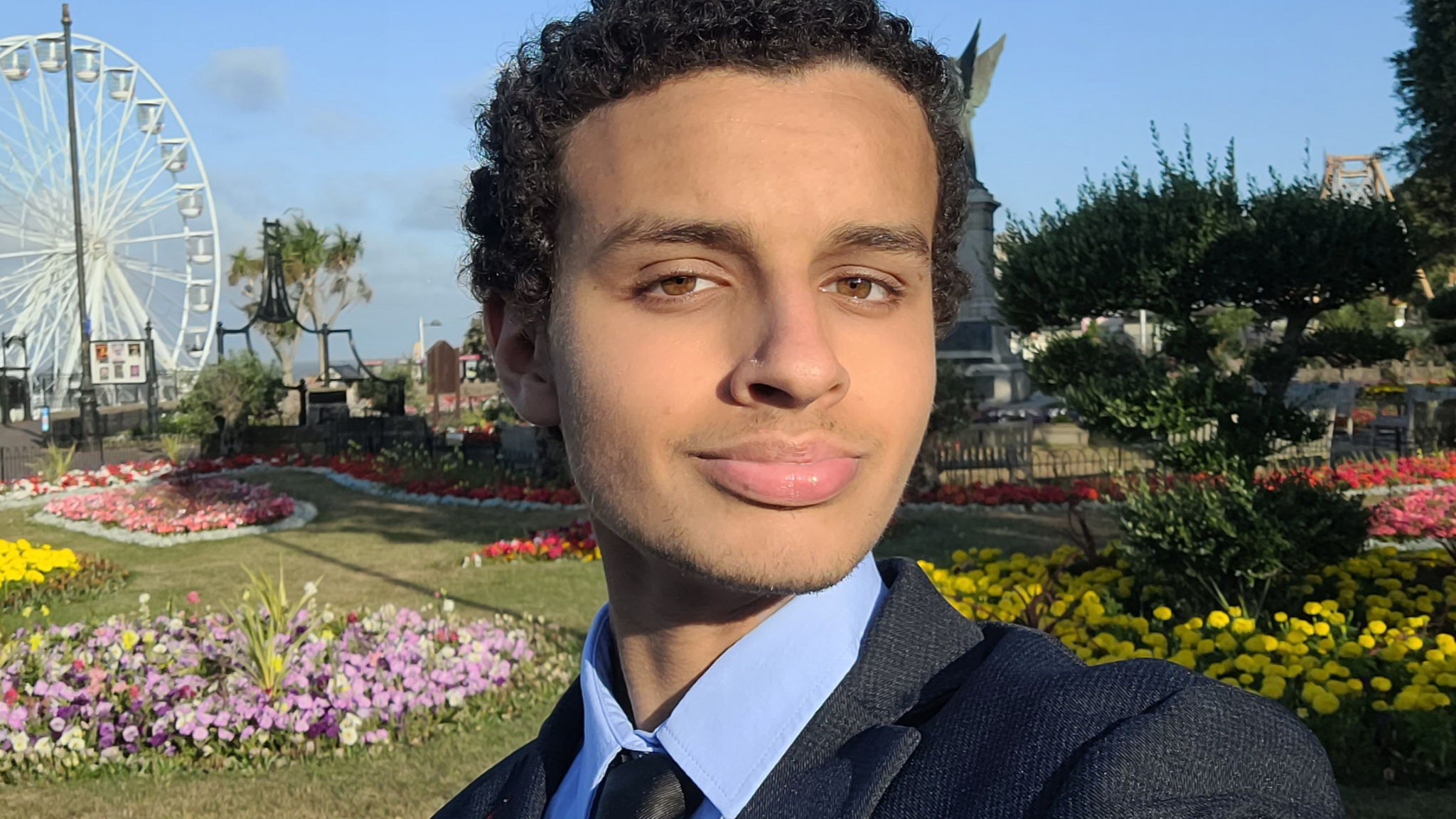 A selfie style picture of Mr Alkhatip who has short curly black hair and is wearing a dark suit with a blue shirt. He is standing in the cliff top gardens in Clacton with a ferris wheel in the background