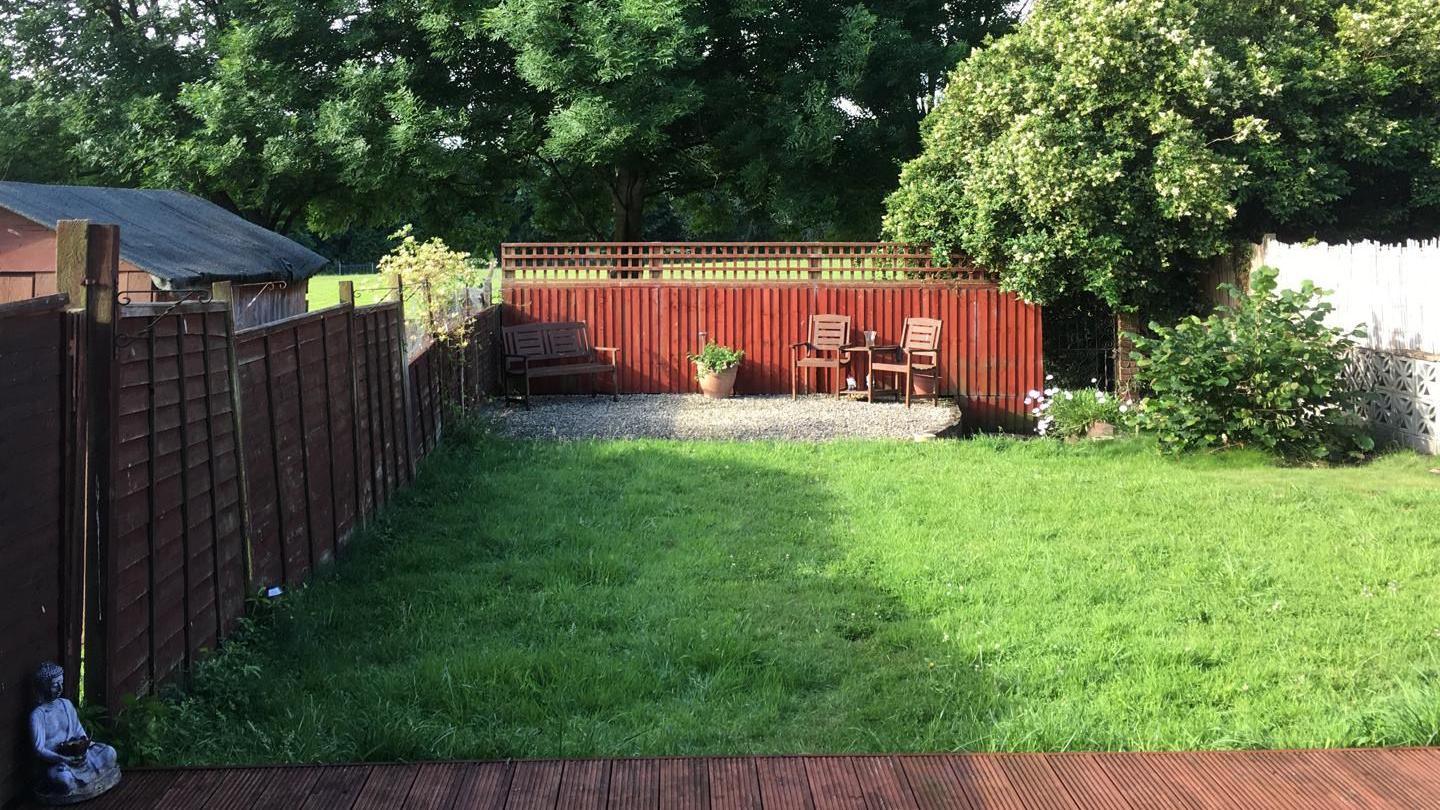 A garden which has decking and leads on to a large lawn. At the back of the garden there is a gravel seating area with two wooden chairs and a wooden bench. Surrounding the garden is a wooden fence. Between the seating is a single plant pot. In the far right corner of the garden there is a bush.