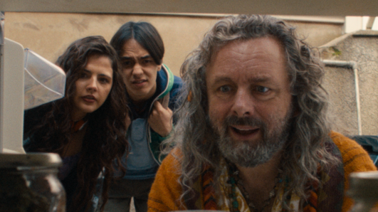 A man with long, curly grey hair and facial hair, staring into a cupboard full of jars. A young man and young woman, both brunette, are stood behind him, also leaning down to look at what he is lookin at. They have puzzled looks on their faces.