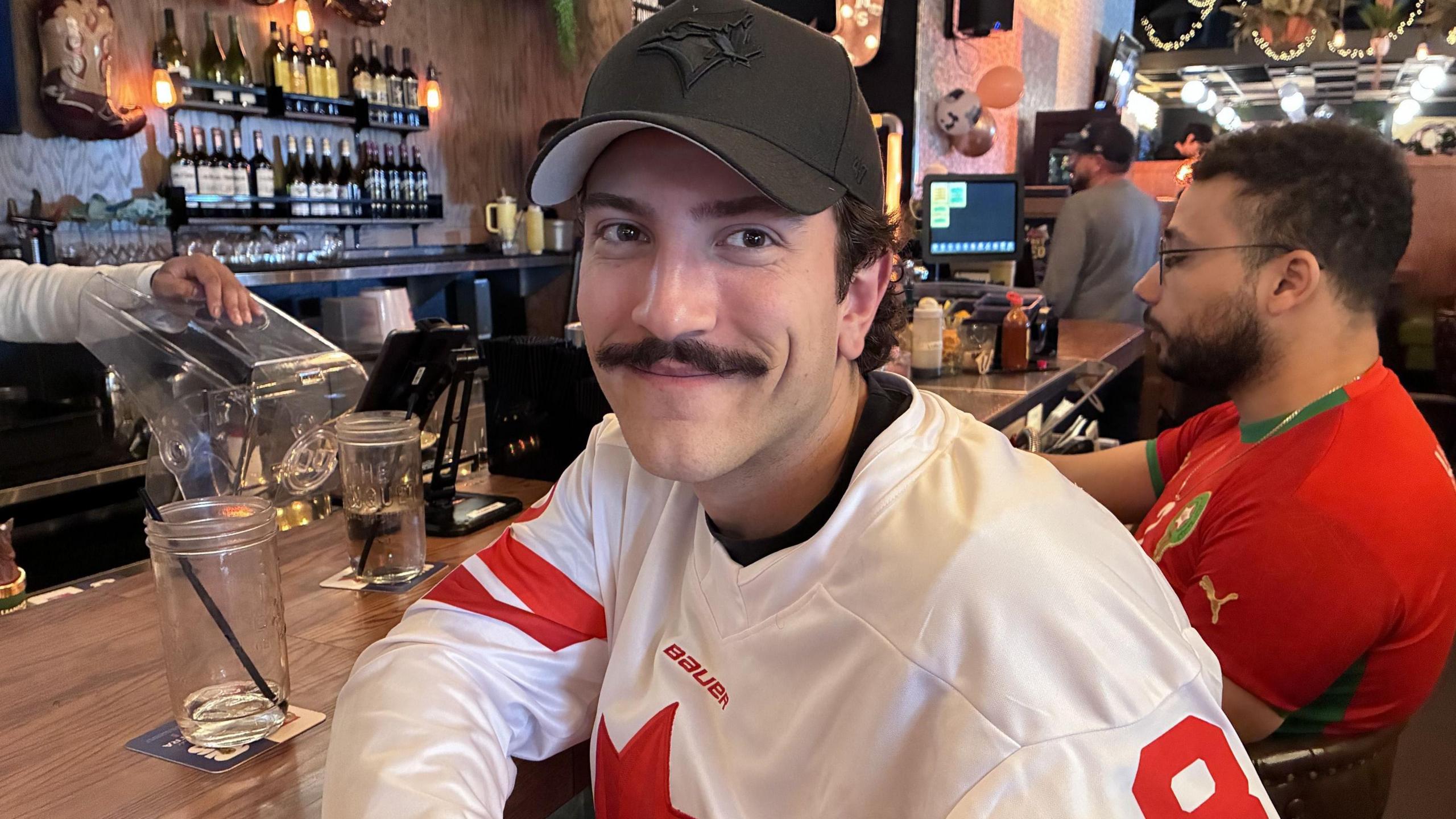 Matt Lorincz, with a moustache,  wears a black Blue Jays baseball cap and a white and red Canada jersey. He is smiling and resting one arm on a bar counter.