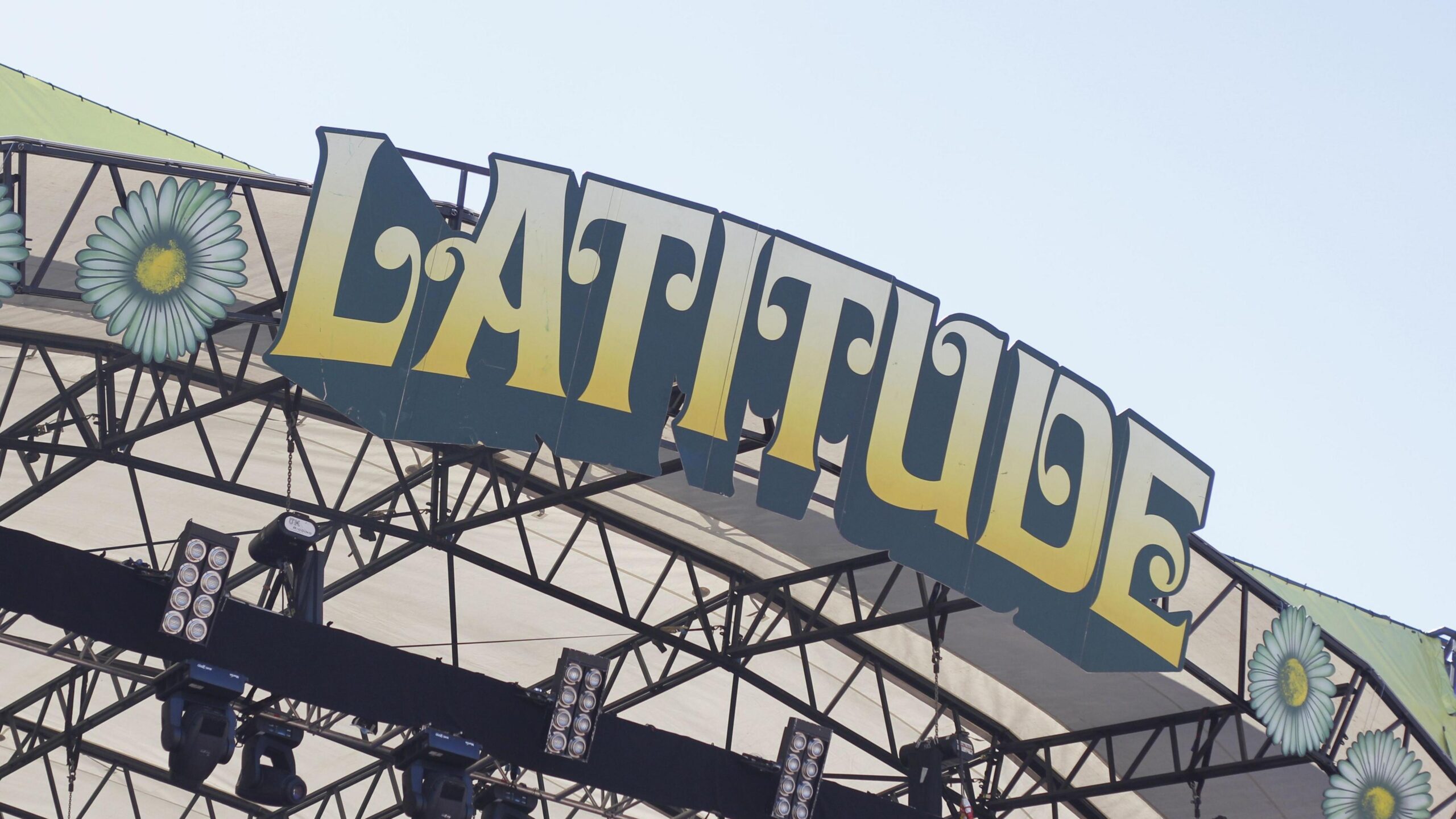 A close-up of the Latitude Festival sign on the top of a stage at the event. The letters are in block capitals, edged in black and coloured yellow fading to cream. Above it is sky.