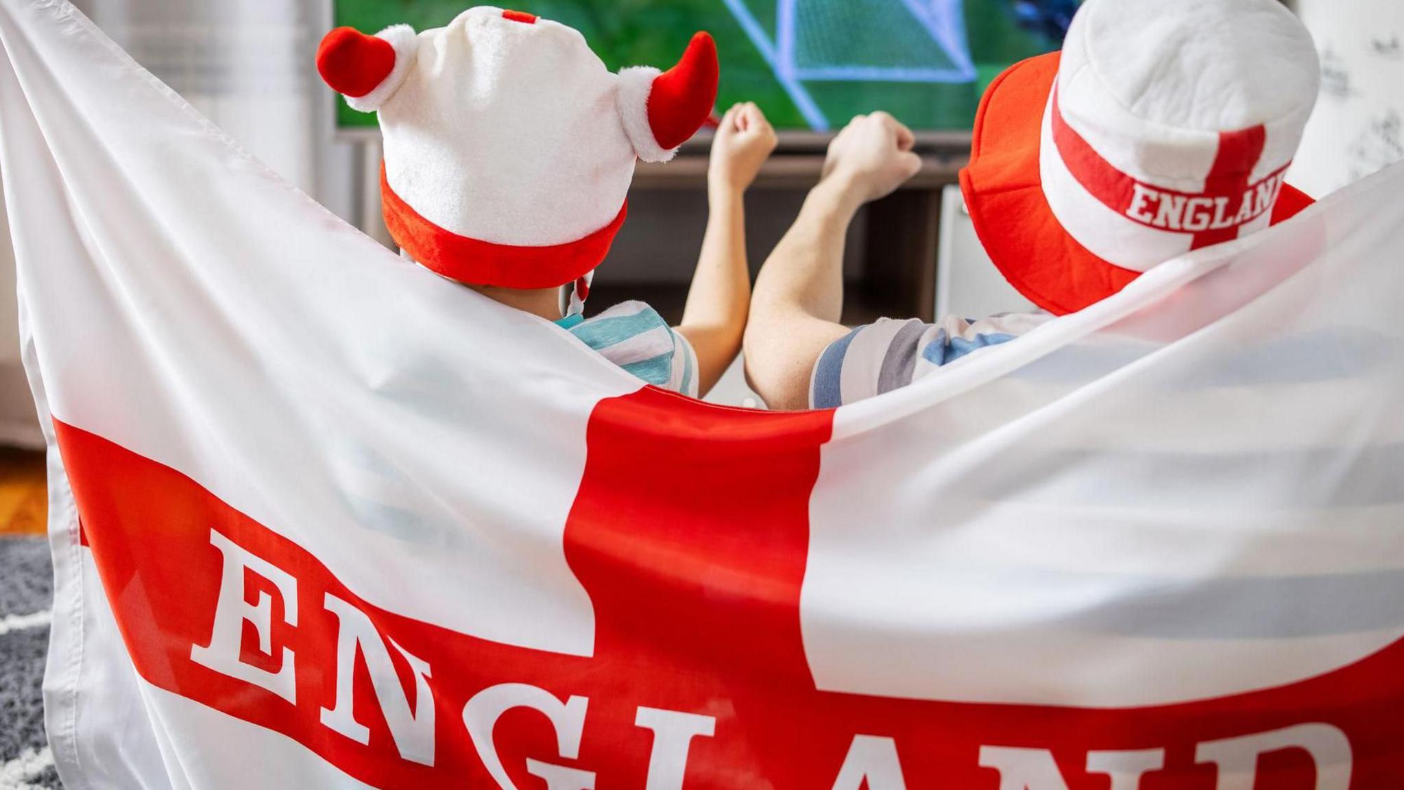 Two young England fans drapped in a ST George's cross cheer as they watch the Three Lions play on tv.