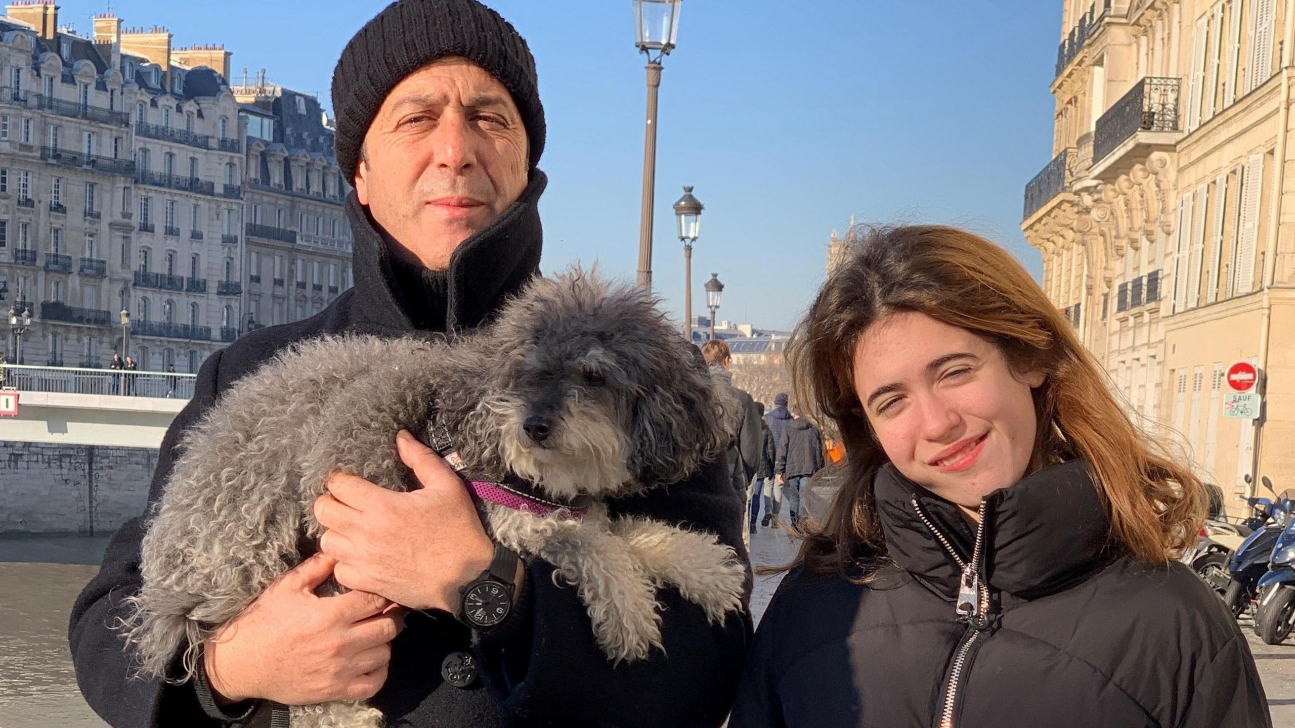 A father and daughter standing by a river in a city. They are wearing warm coats, and the father is holding a fluffy grey dog with a pink lead.