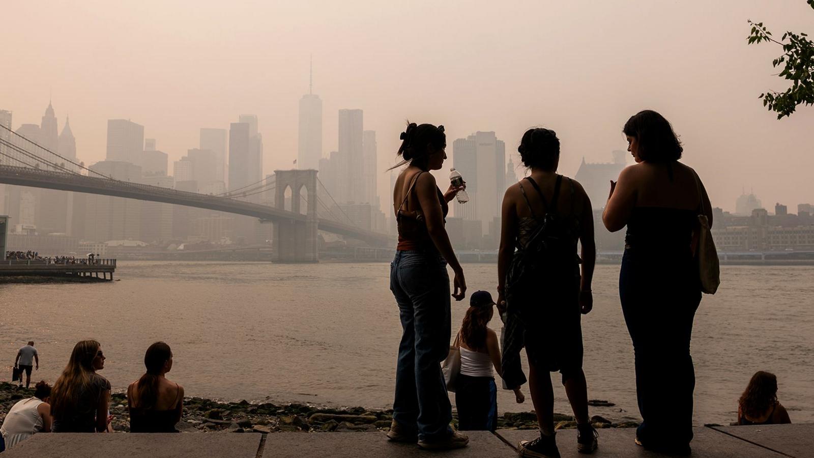 People are silhouetted against the New York City skyline with Brooklyn bridge and the river in view, and a hazy smokey sky, in Brooklyn on 16 July.