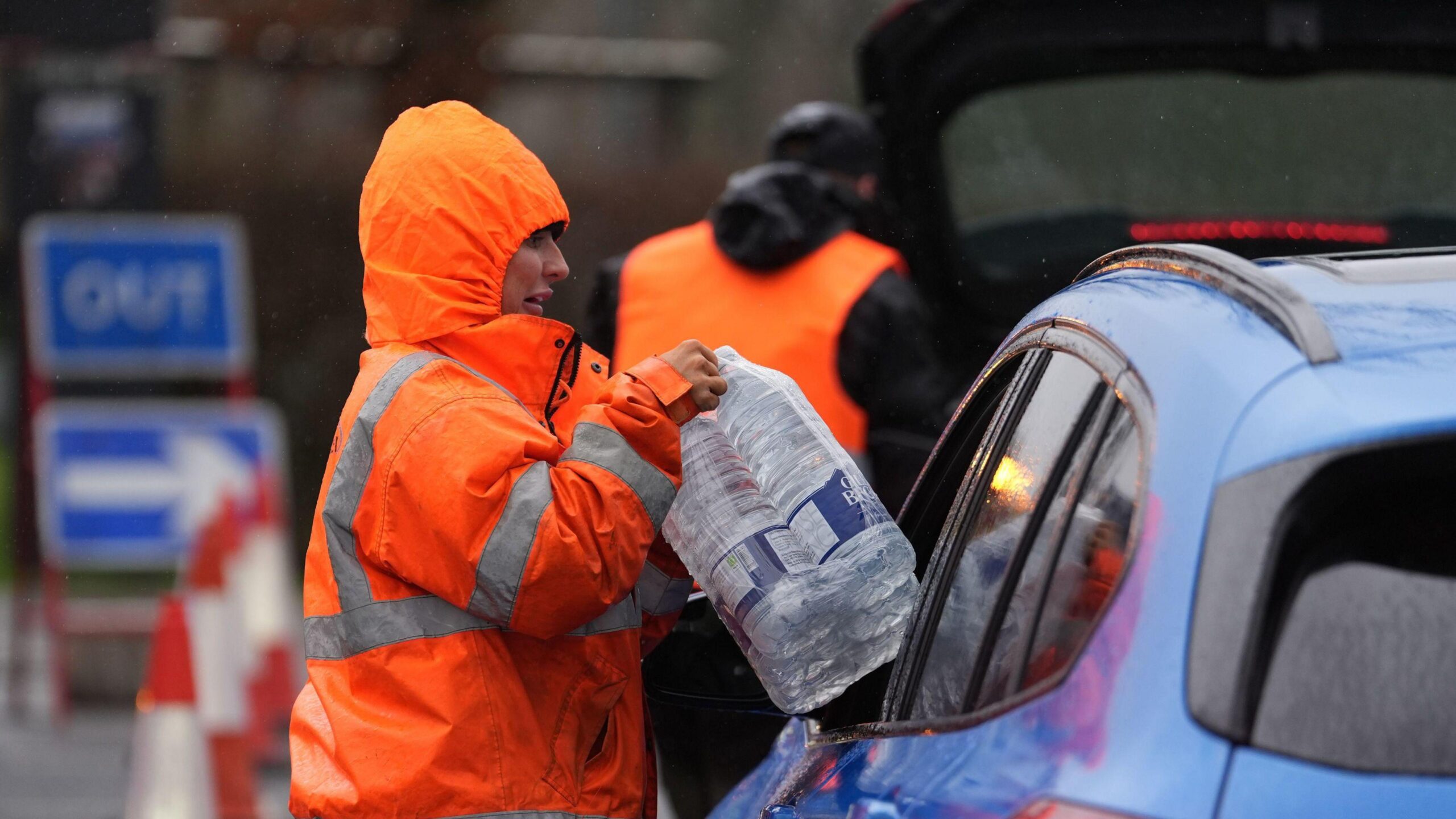 A woman in an orange jacket hands bottles of water through a car window.