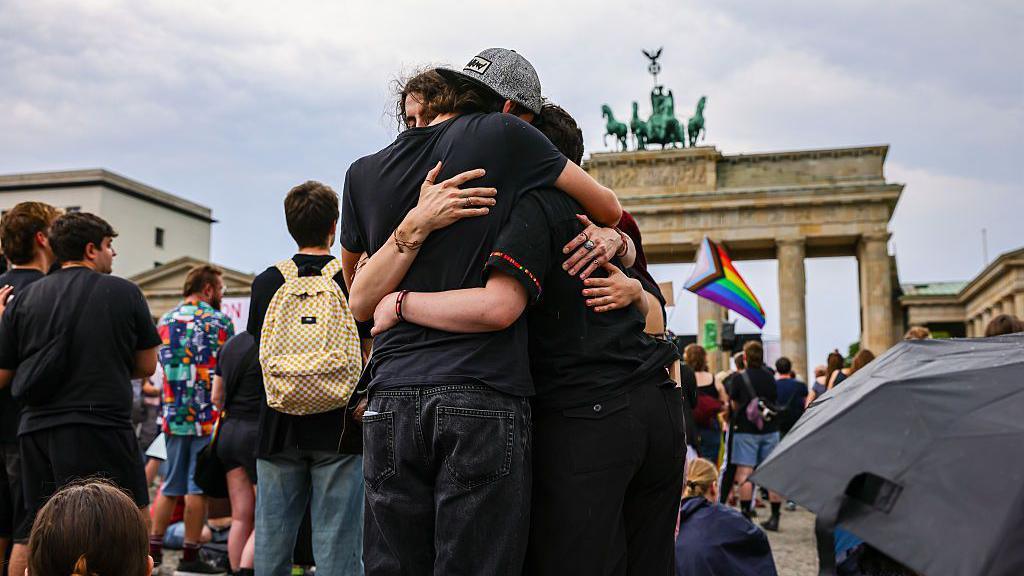 Three people hug each other with a rainbow LGBT flag visible in the background and the Brandenburg Gate behind