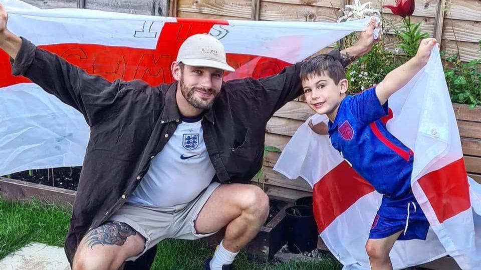 Daniel Beames, wearing an England shirt and dark shirt, and his partner's son, Barney, wearing a blue England shirt, both hold England flags aloft in a garden in Bishop's Cleeve during the Euros in 2021.