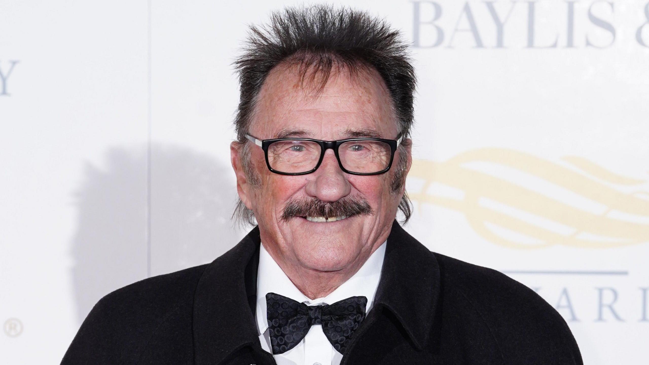 Paul Chuckle at a red carpet event. It is a close crop of his face. He has a brown moustache, brown hair sticking up around his head and black square glasses. He is smiling and wears a white shirt, black bowtie and black jacket.