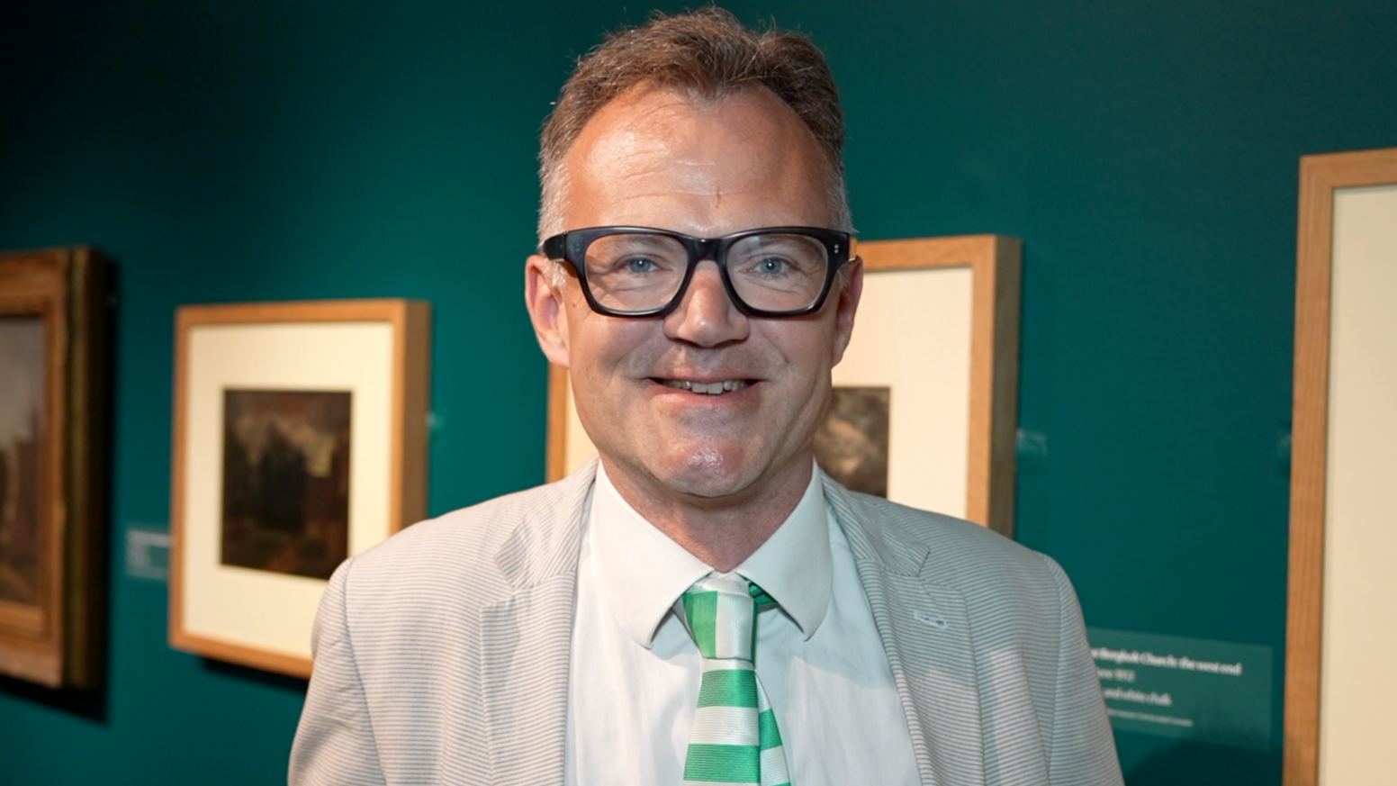 A man smiles at the camera at an art exhibition. He has short grey hair and is wearing a light grey suit, glasses, white shirt and a green and white tie. Pieces of art are on the wall behind him.