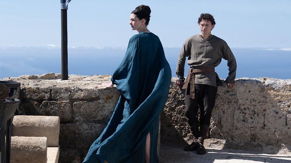 Anne Hathaway as Penelope (Odysseus's wife) in centre of image in blue gown looking to the side and Tom Holland as their son Telemachus leaning on wall in khaki top and dark trousers. Blue skies behind
