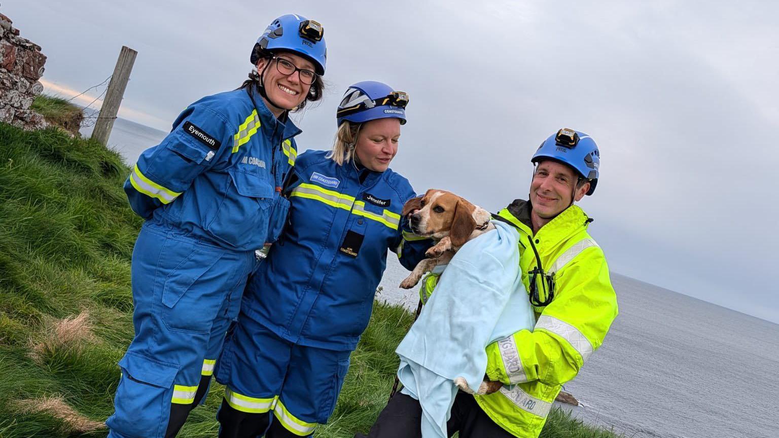 Two people in blue outdoor wear beside a man in a high vis jacket who is holding a dog wrapped in a blanket