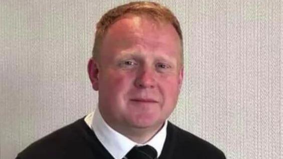Robert has receding ginger hair and is red-faced. He is wearing a black jumper over a white shirt and black tie.