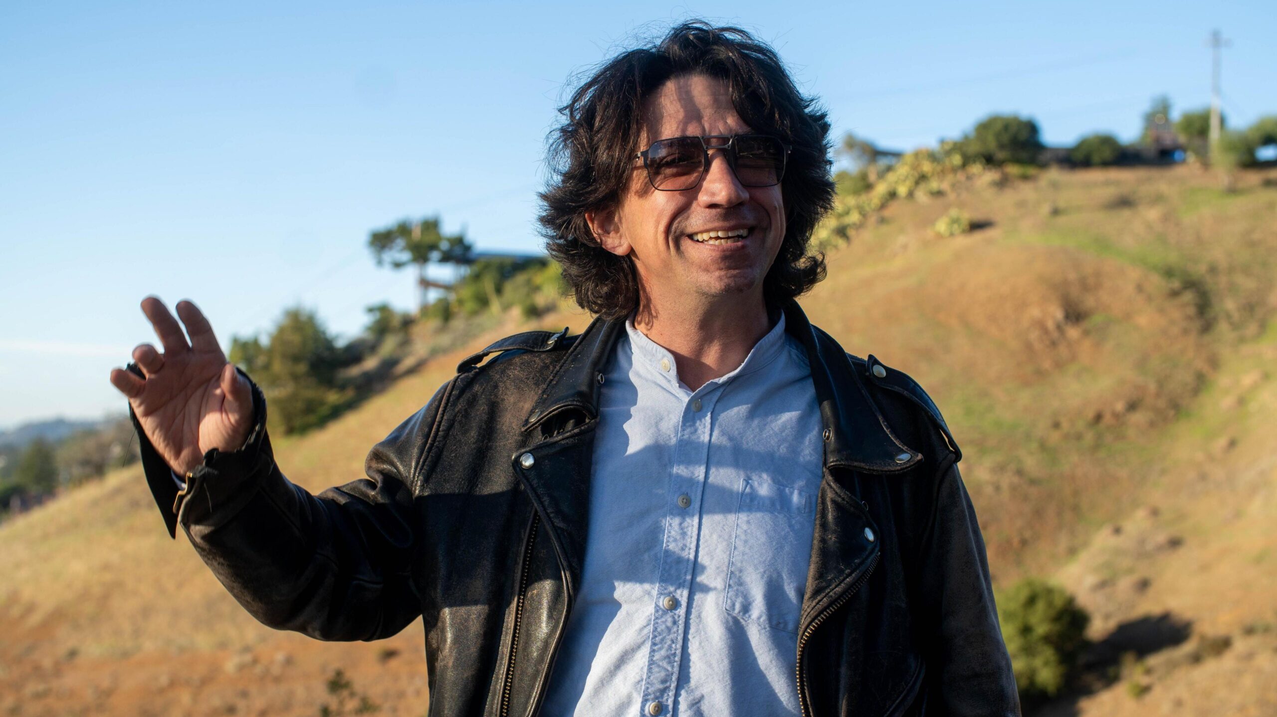 A man in a leather jacket and shades, with long dark hair, smiles and raises his right hand as he stands in front of a sun-baked hillside