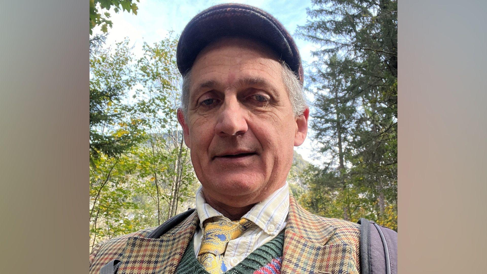 A selfie-style picture of Robert Elwes, who is wearing a flat cap, checked jacket and a multi-coloured jumper with yellow and blue tie. He is standing outside surrounded by trees.