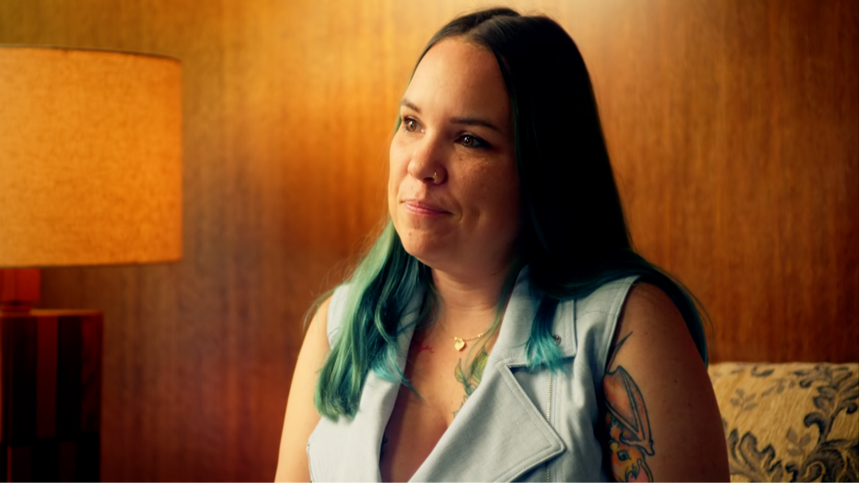 Taylor, a woman with long, dark hair and a tattoo over part of her arm and her upper chest, wears a pale green sleeveless top and sits in a room with wood-panelled walls and an orange lamp.