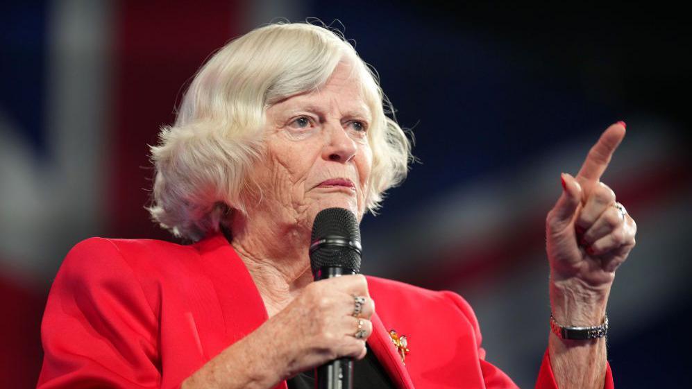 Ann Widdecombe speaking at the Reform UK party conference in 2024. She has a bob style haircut with mostly white hair, is wearing a red blazer with a broach, and is holding a microphone in her right hand as she speaks, while making a pointing gesture with her left hand.