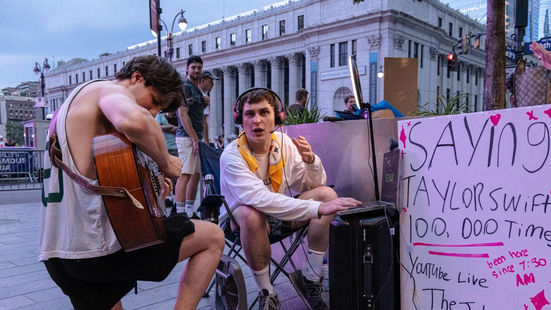 A man playing a guitar kneels next to a man sat in a chair alongside a sign that reads: "Saying 'Taylor Swift' 100,000 times".