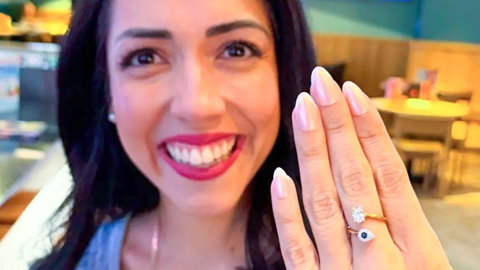 A close up of a young woman smiling broadly and holding her hand up in front of her. She has a ring on her middle finger with a sapphire and diamond.