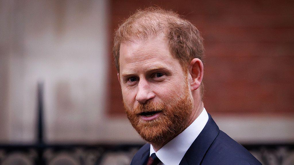 Headshot of Prince Harry, who has red hair and a beard, looking off-camera wearing a navy blue suit with white shirt, and navy blue and maroon-striped tie.