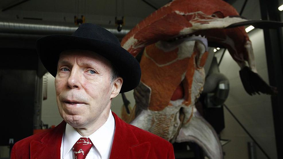 Gunther von Hagens, wearing a red jacket and tie with white shirt, and a black hat, stands in front of an exhibit in Guben, Germany in 2016.
