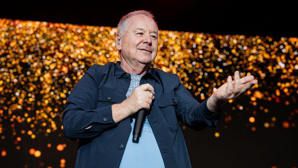 A man with short grey hair is holding a microphone on stage