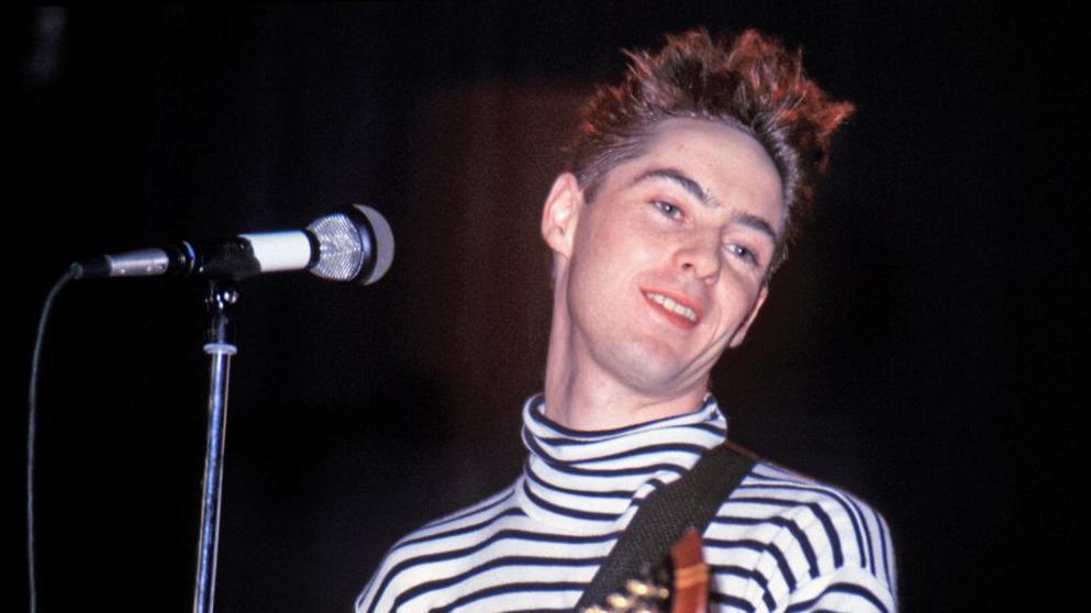 Roddy Frame performing on stage in the eighties. He is wearing a black and white polo top. He is in front of  mic and has a guitar over his shoulder