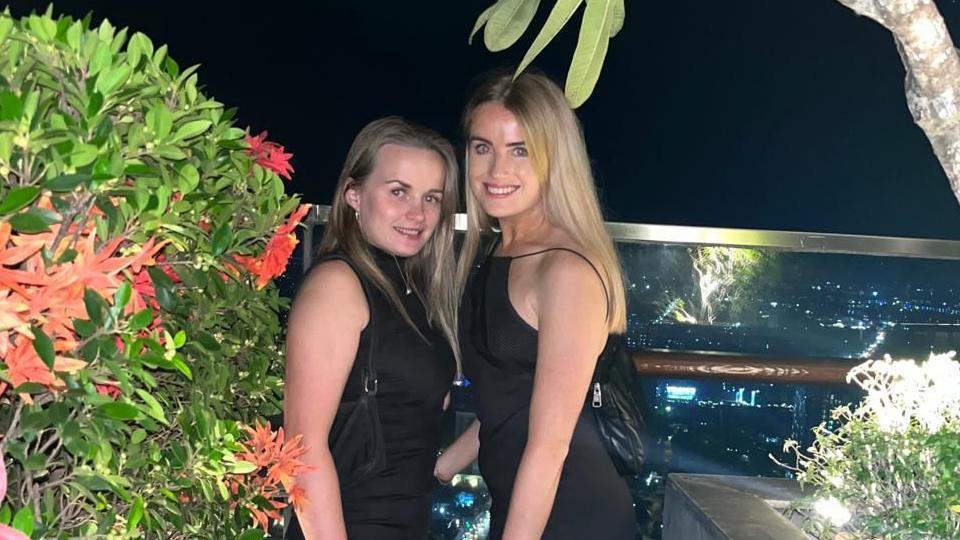 Two women, wearing all-black dresses, posing on of a balcony at night, surrounded by some plants. They're both smiling at the camera.