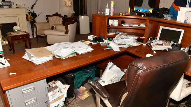 A pristine-looking area with armchairs and flowers contrasts with a messy desk. Under the desk, hidden from view, are various papers and carrier bags.