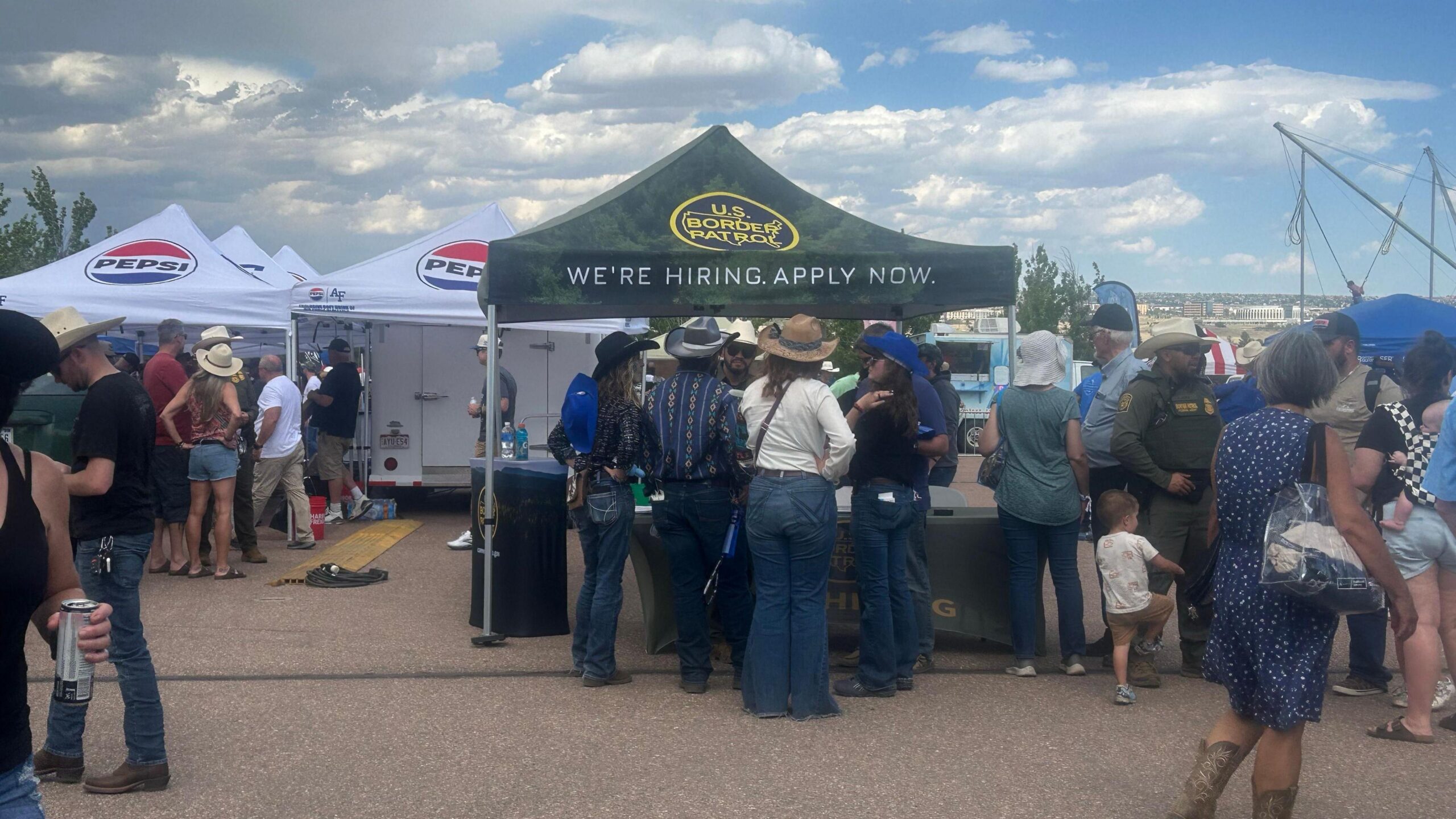 Folks wearing cowboy hats and jeans line up in front of a tent emblazoned with the Border Patrol logo that reads "We're hiring. Apply now"