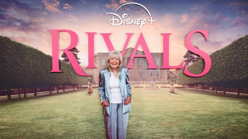 Dame Jilly Cooper wearing a blue suit and trousers. She stands in front of a "Rivals" sign as she attends a special UK screening of the show