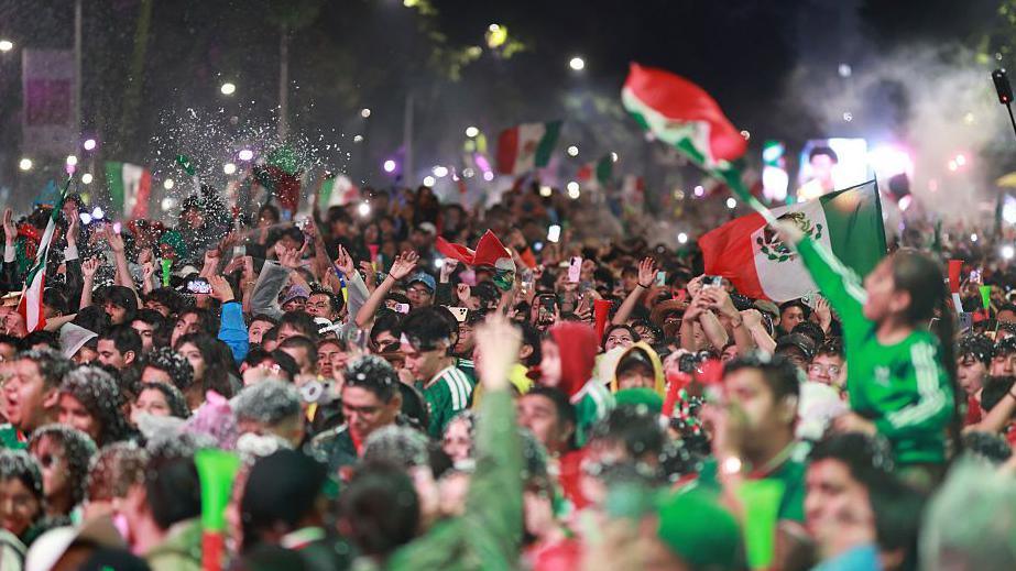 A huge crowd of fans wearing Mexico's green football kit and waving the country's flag celebrate the team's 2-0 World Cup win against Ecuador in Mexico City at night.