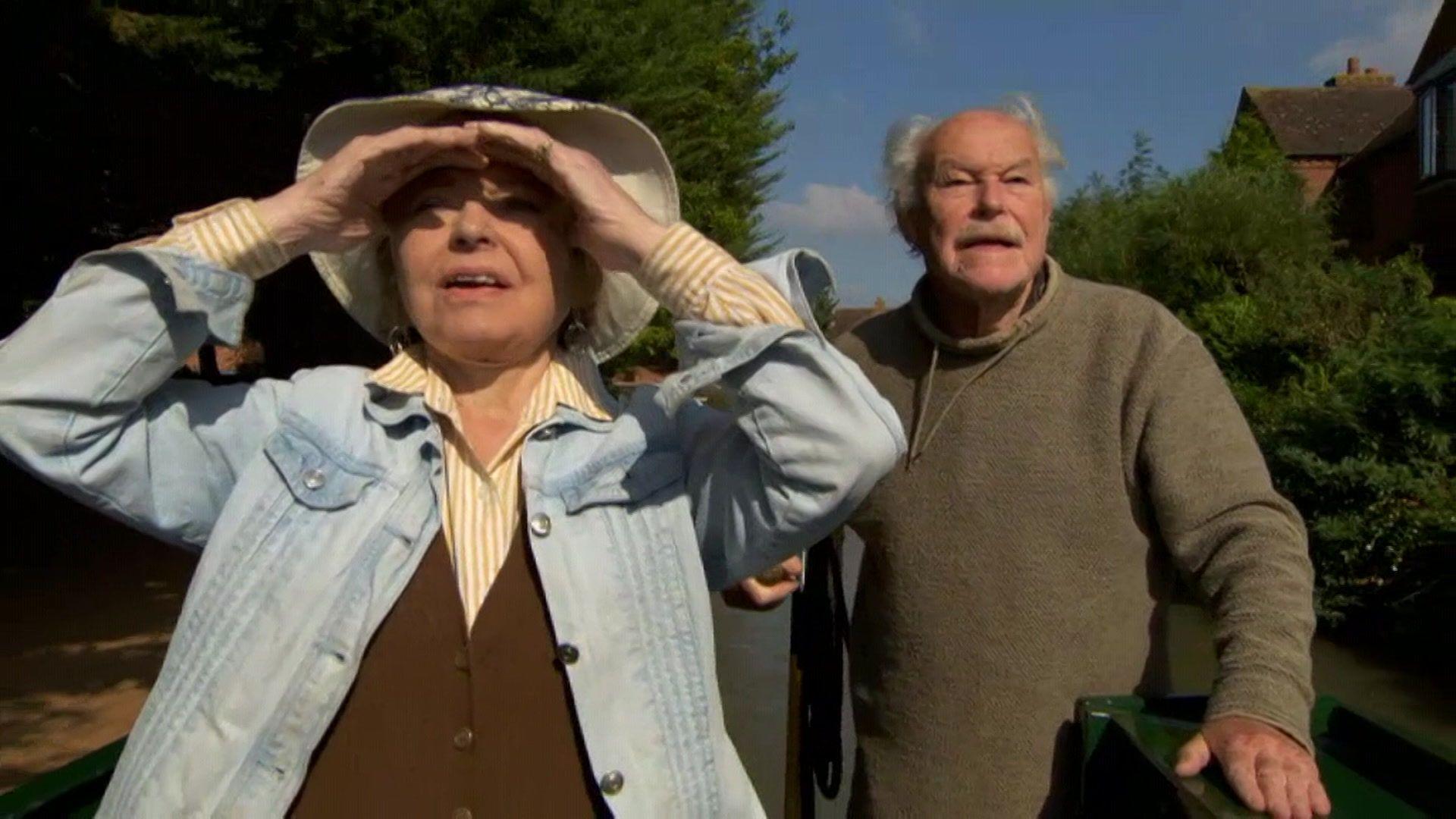 Prunella Scales and Timothy West on a canal boat. Timothy is in brown jumper and looking off in the distance. Prunella is in a yellow shirt, brown waistcoat with a blue denim jacket. She is also looking in to the distance but shielding her eyes from the sun.