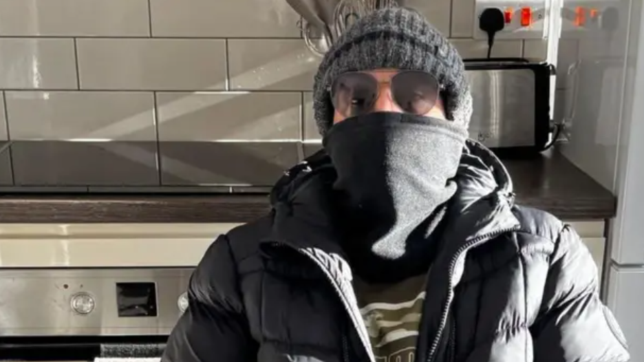 A man wearing dark glasses, a puffer jacket, scarf around his face and beanie hat is sitting in a kitchen.