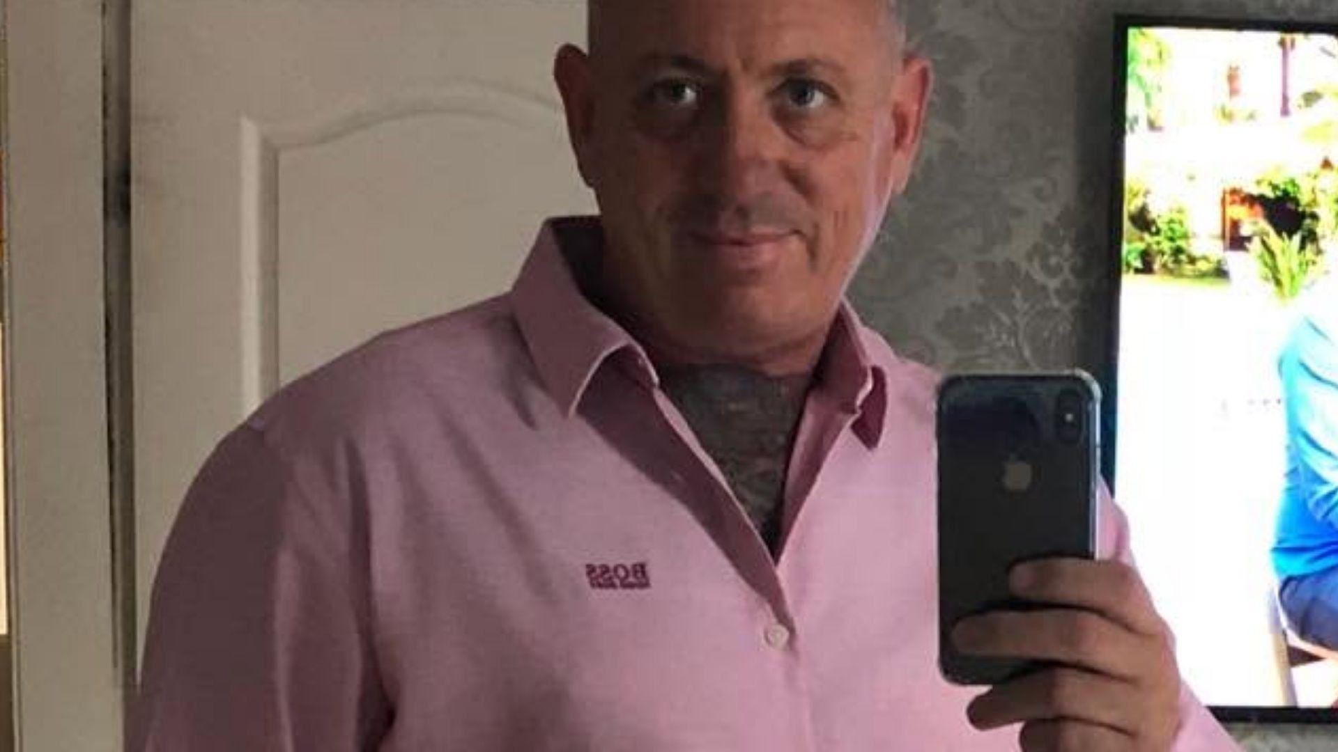 A middle aged man takes a selfie with his mobile phone in a bedroom mirror. He is wearing a pink Hugo Boss branded buttoned shirt with an open collar, has short grey hair and the front of his chest up to his neck is covered in tattoos.