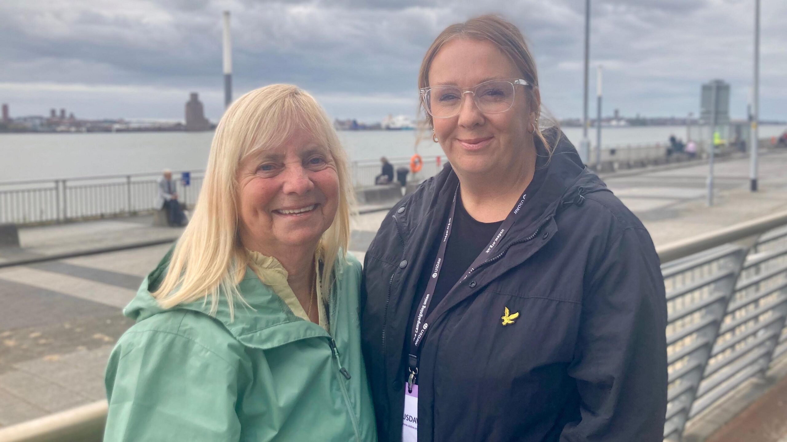 Two women stand by the river Mersey. One is blonde and wears a pale green light anorak. The other women is taller, with long hair tied into a ponytail. She has glasses and is wearing a navy blue jacket. They are both smiling at the camera.